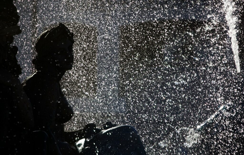 A classical statue silhouetted against glittering, backlit fountain spray