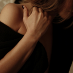Woman in a black off-the-shoulder sweater holding the back of her neck, face turned away, in dim warm light.