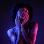 Close-up of a woman with curly hair, eyes closed, holding both hands to her cheeks under blue and pink lighting.